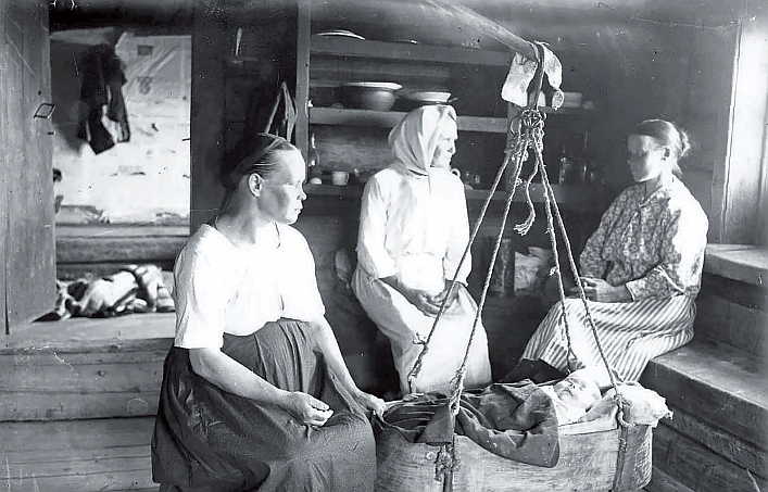 Mustavalkoinen kuva kolmesta naisesta mökissä kehdon äärellä