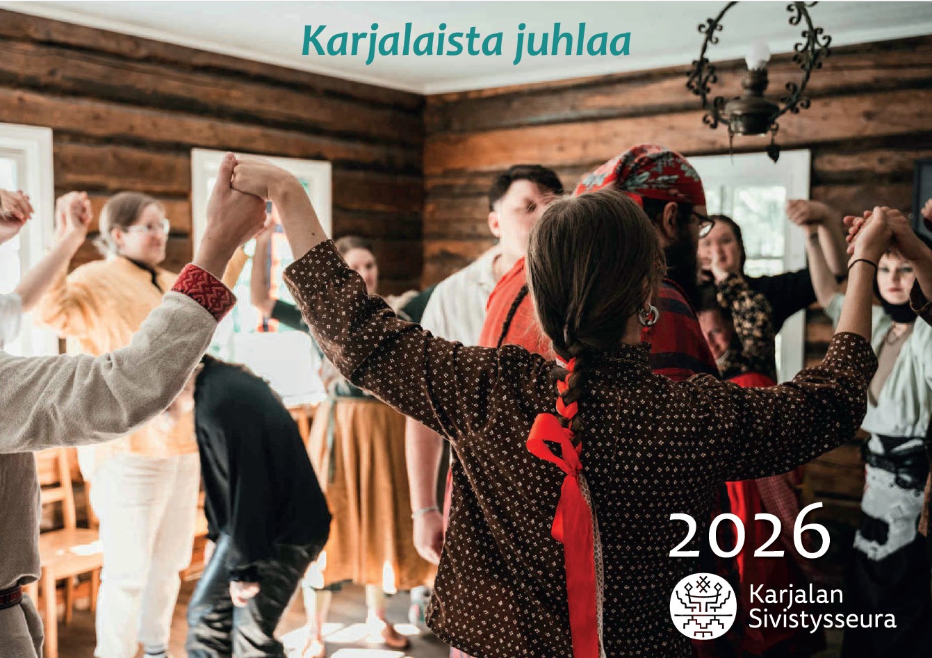 Kalenterikansi_2026_Karjalan-Sivistysseura Hirsiseinäisessä pirtissä on kuvattu läheltä piirissä tanssivaa ryhmää. Perinteisiin karjalaisasuihin pukeutuneet ihmiset pitävät toisiaan kädestä kiinni, kädet kohotettuina ylöspäin kohti kattoa. Kuvan yläreunassa on teksti "Karjalaista juhlaa" ja oikeassa alakulmassa teksti 2026, Karjalan Sivistysseura.