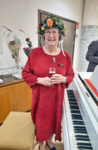 Seppelepäinen, punaiseen pellavamekkoon pukeutunut nainen hymyilee kameralle kädessään kuohuviinilasi, vierellään valkoinen flyygeli, taustallaan korkeita kukkakimppuja ja päässään muhkea kukkaseppele.