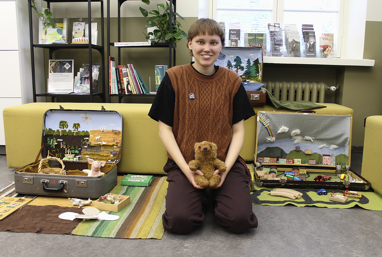 Lattialla, polviensa päällä istuu hymyilevä nuori, jonka sylissä on pieni ruskea nalle ja hänen vierellään kahta puolen on avoimia matkalaukkuja, joissa on värikkäitä, nukkekotimaisia maailmoja. Taustalla on ikkuna ja kirjahylly, lattialla molempien matkalaukkujen alla värikäs matto ja leluja, esimerkiksi Brion puisen junaradan kappaleita.